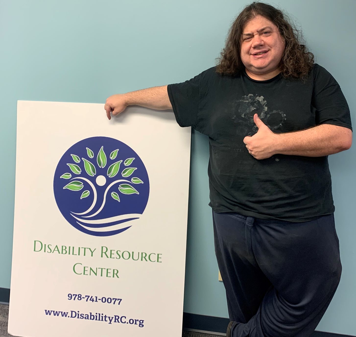 A white man with long curly brown hair leaning on a Disability Resource Center poster giving a thumbs up and a smile