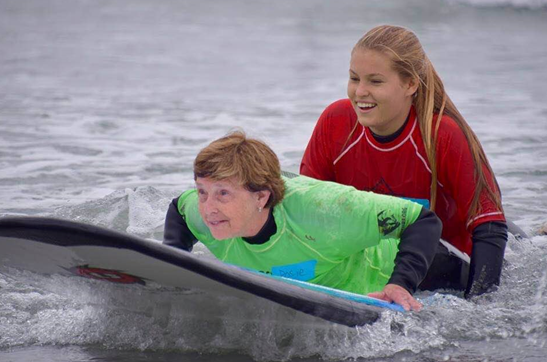 Image of a woman with a green shirt getting help from another woman with a red shirt to surf in the water